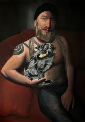 A shirtless, bearded man with a large scarrification/tattoo on his shoulder, holding a long haired merle chihuahua