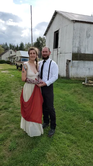 Stephanie in her wedding dress with her husband. Dress is comprised of an ecru linen bodice, with ecru silk chiffon skirt and red silk chiffon overdrape. A wool and silk band joins the bodice to the dress. Styled with wool arm warmers covered in white leaf pattern.
